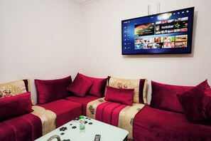 Ein Wohnzimmer mit einem großen Flachbildfernseher an der Wand, der eine Streaming-Oberfläche anzeigt. Ein rotes Sofa mit passenden Kissen und einer gemusterten Decke ist ebenfalls zu sehen. Im Vordergrund steht ein kleiner Tisch mit einer Fernbedienung und einer Flasche.