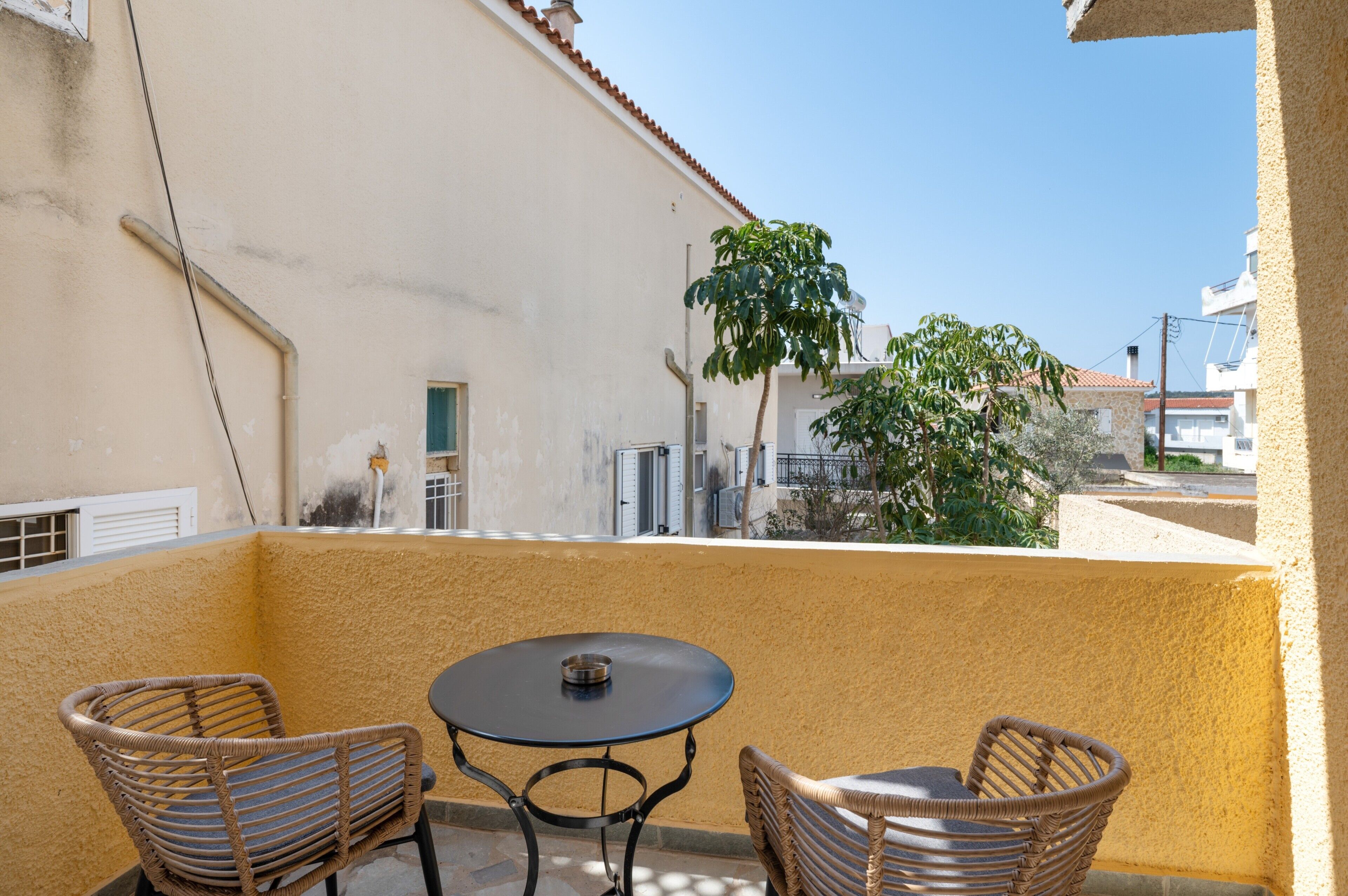 Un balcon avec deux fauteuils en osier et une petite table ronde, d’où l’on peut admirer un immeuble avec un toit en tuiles et un arbre.