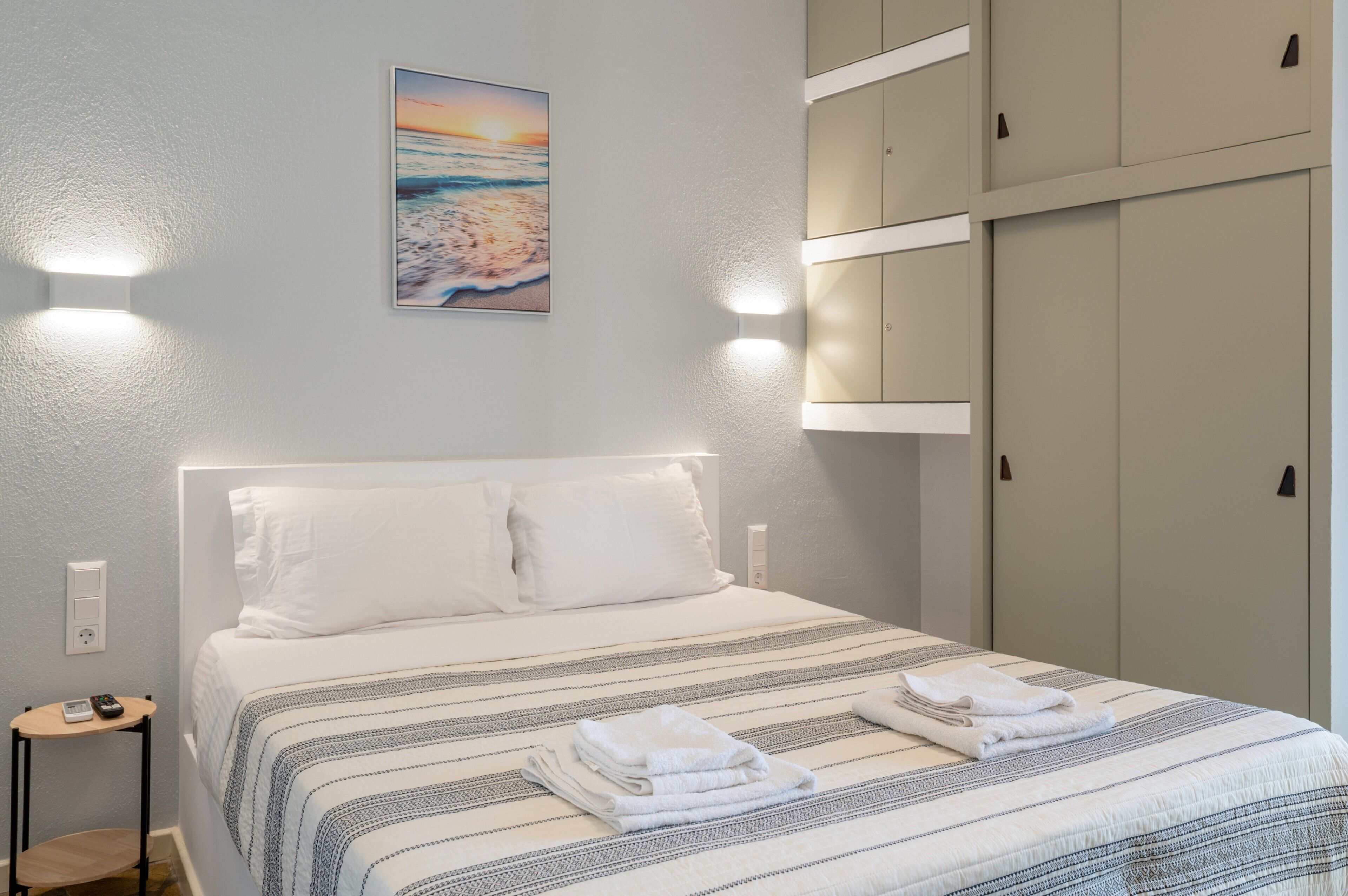 Een netjes opgemaakt bed met een gestreept beddegoed, een aan de muur hangend schilderij van een zonsondergang, en een aantal kasten aan de rechterkant.