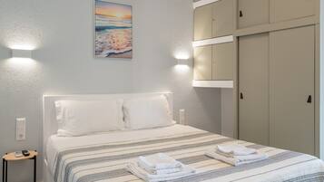 Een netjes opgemaakt bed met een gestreept beddegoed, een aan de muur hangend schilderij van een zonsondergang, en een aantal kasten aan de rechterkant.