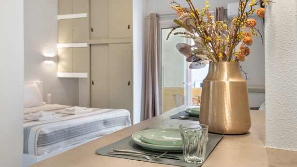 Una habitación de hotel con una cama bien hecha, una mesa de comedor con platos y cubiertos, y un jarrón con flores sobre la mesa.