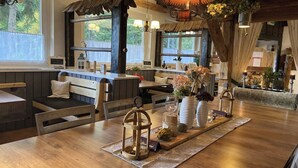 Un restaurante acogedor con muebles de madera, una mesa central adornada con velas y flores, y grandes ventanales que ofrecen vistas al verdor.