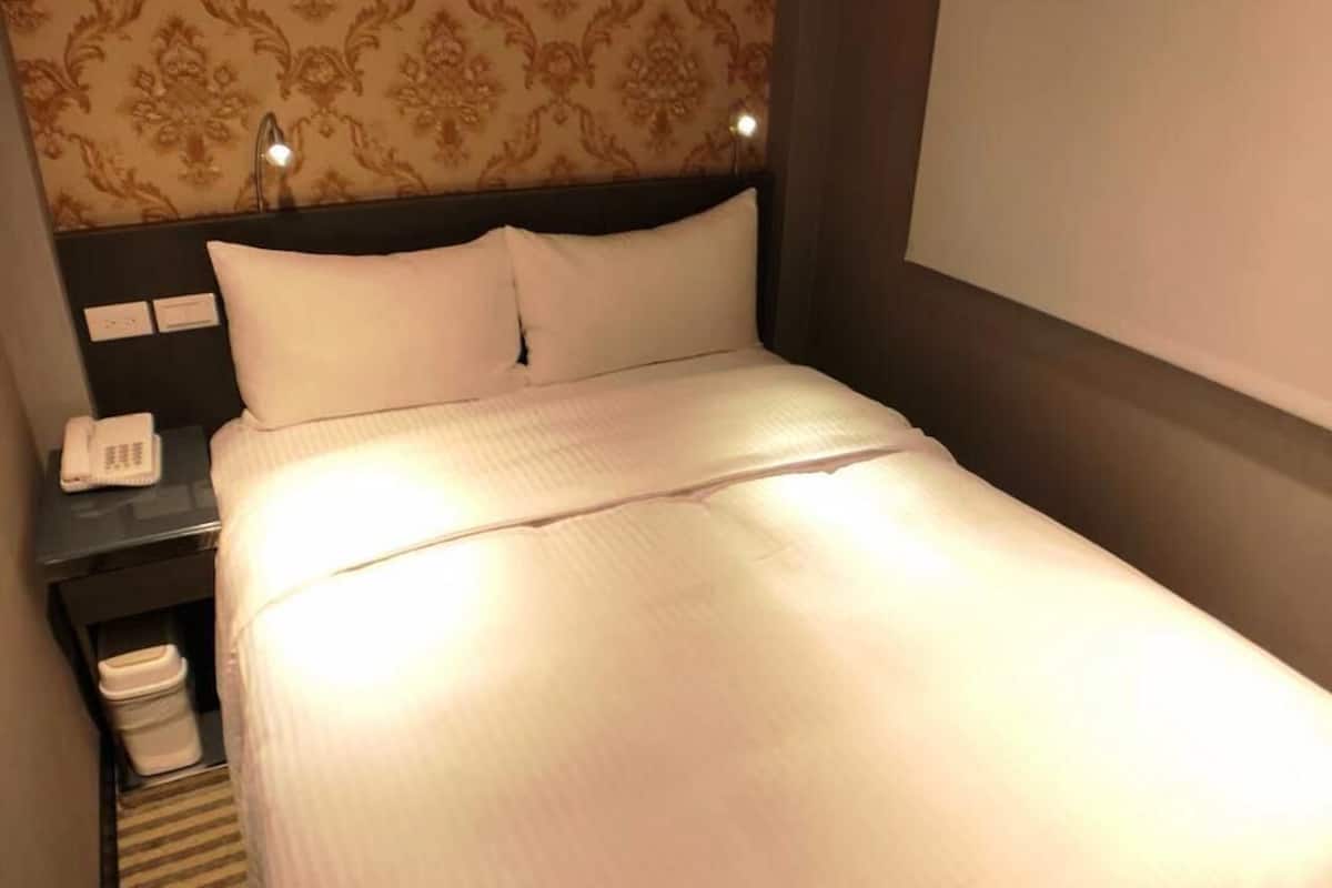 A hotel room with a neatly made bed, a nightstand with a phone, and a patterned wallpaper.