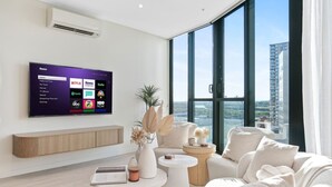 A modern living room with a large flat-screen TV displaying streaming services, a comfortable seating area with a sofa and armchairs, a coffee table, and a view of a cityscape through large windows. - Bouclé Haven | 2BR, Sunset Skyline Views + Parking (Wentworth Point)