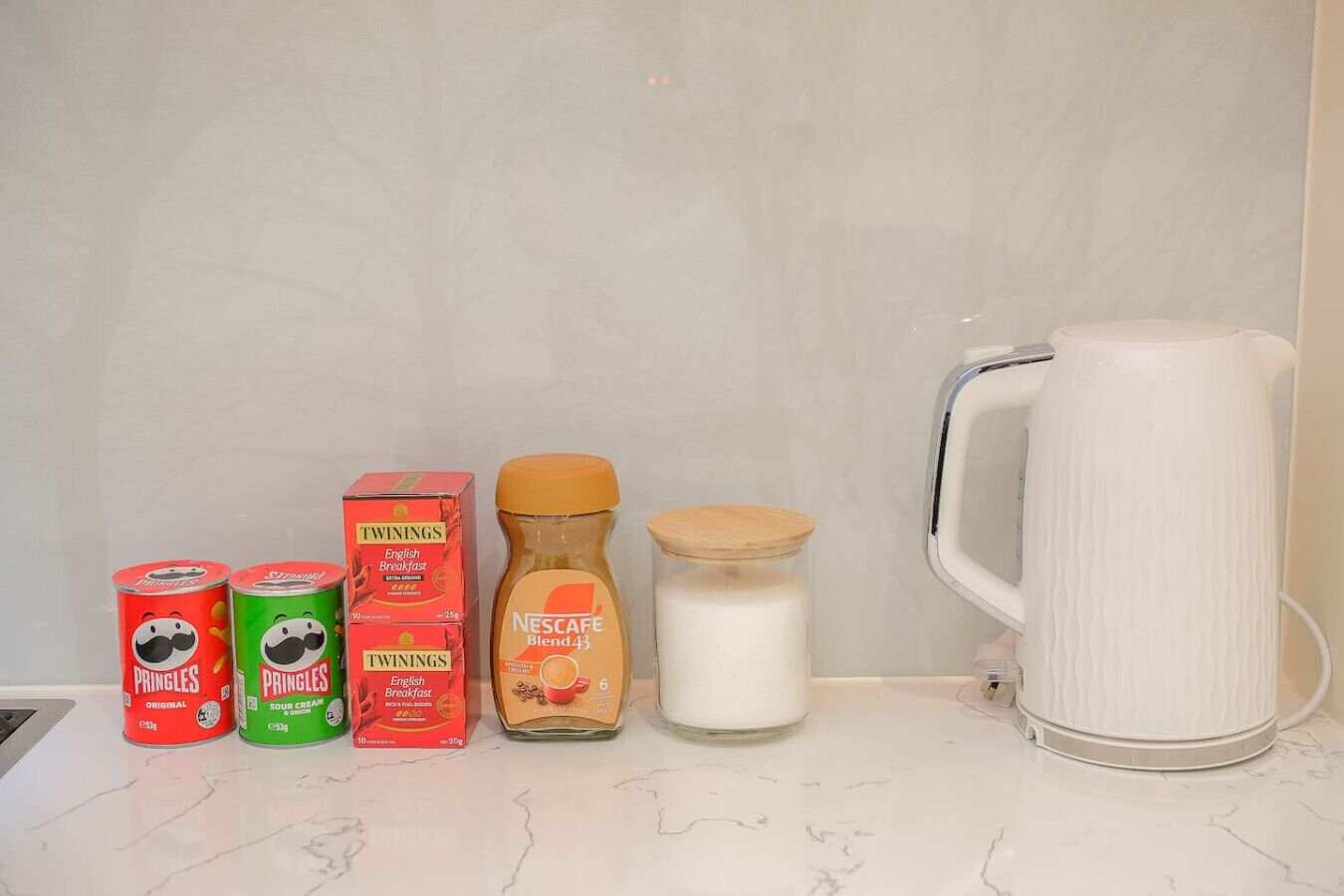 A kitchen counter with a white marble surface, containing two cans of Pringles, a box of Twinings English Breakfast tea, a bottle of Nescafe Blend 43, a jar of milk, and a white ceramic mug.
