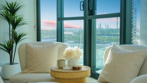 A modern living room with two plush armchairs, a round wooden side table, and a potted plant. A large window offers a view of a city skyline. - Bouclé Haven | 2BR, Sunset Skyline Views + Parking (Wentworth Point)
