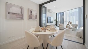A modern dining area with a round table, white chairs, and a view of a cityscape through large windows. - Bouclé Haven | 2BR, Sunset Skyline Views + Parking (Wentworth Point)