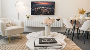 Een moderne woonkamer met een ronde marmeren salontafel, twee witte fauteuils, een witte radiator, een schilderij van een stadsgezicht, en een eethoek met een witte tafel en stoelen.