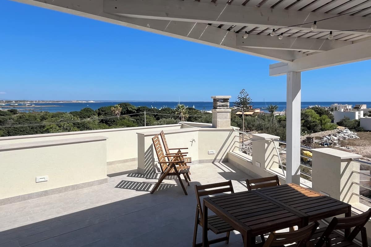Une terrasse avec une table et des chaises en bois, une cheminée en pierre et une vue sur la mer et les bâtiments.