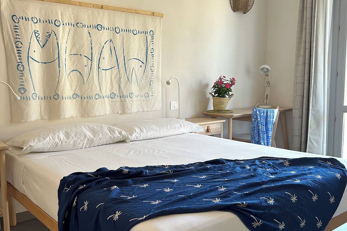 Un lit bien fait, recouvert d’une couverture bleue, une table de chevet avec une lampe, une tapisserie murale et une fenêtre avec des rideaux.