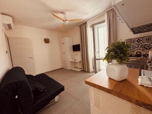 Un ambiente compatto con ventilatore a soffitto, angolo cottura, zona pranzo e piccolo salotto con divano.