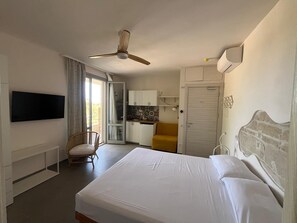 Habitació d'hotel amb llit, televisió, ventilador de sostre, cadira, porta corredissa a un balcó i una cuineta petita.