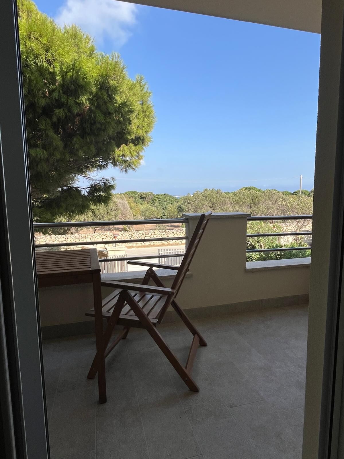 Un balcon avec une chaise en bois et une table, donnant sur un paysage côtier avec des arbres et un ciel dégagé.