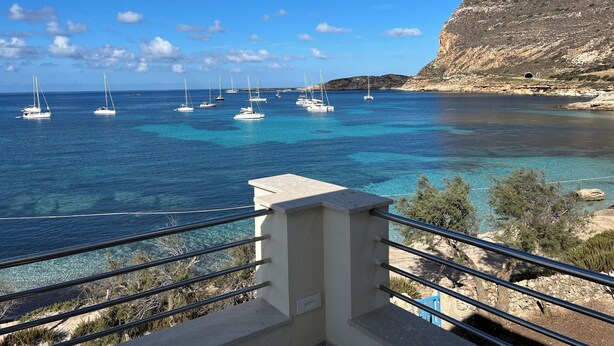 Vista sulla costa con diverse barche a vela ancorate in acque blu cristalline, una scogliera rocciosa e una ringhiera da balcone.