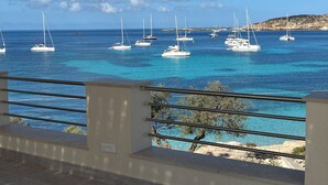 Vista sulla costa con diverse barche a vela ancorate in acque blu cristalline, una linea di costa rocciosa e una ringhiera da balcone in primo piano.