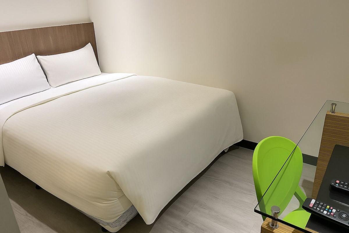 Et hotelværelse med en stor seng, en grøn lænestol og et skrivebord med en computer og fjernbetjeninger.