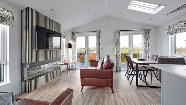 A spacious living area with a large flat-screen TV, a fireplace, a sofa, a dining table with chairs, and a view through glass doors.