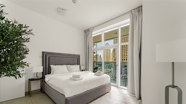 A modern hotel room with a large bed, a bedside table with a lamp, a potted plant, and a view of the city through large windows. - Stylish 2BR in Dubai Marina Near the Beach (Dubai)