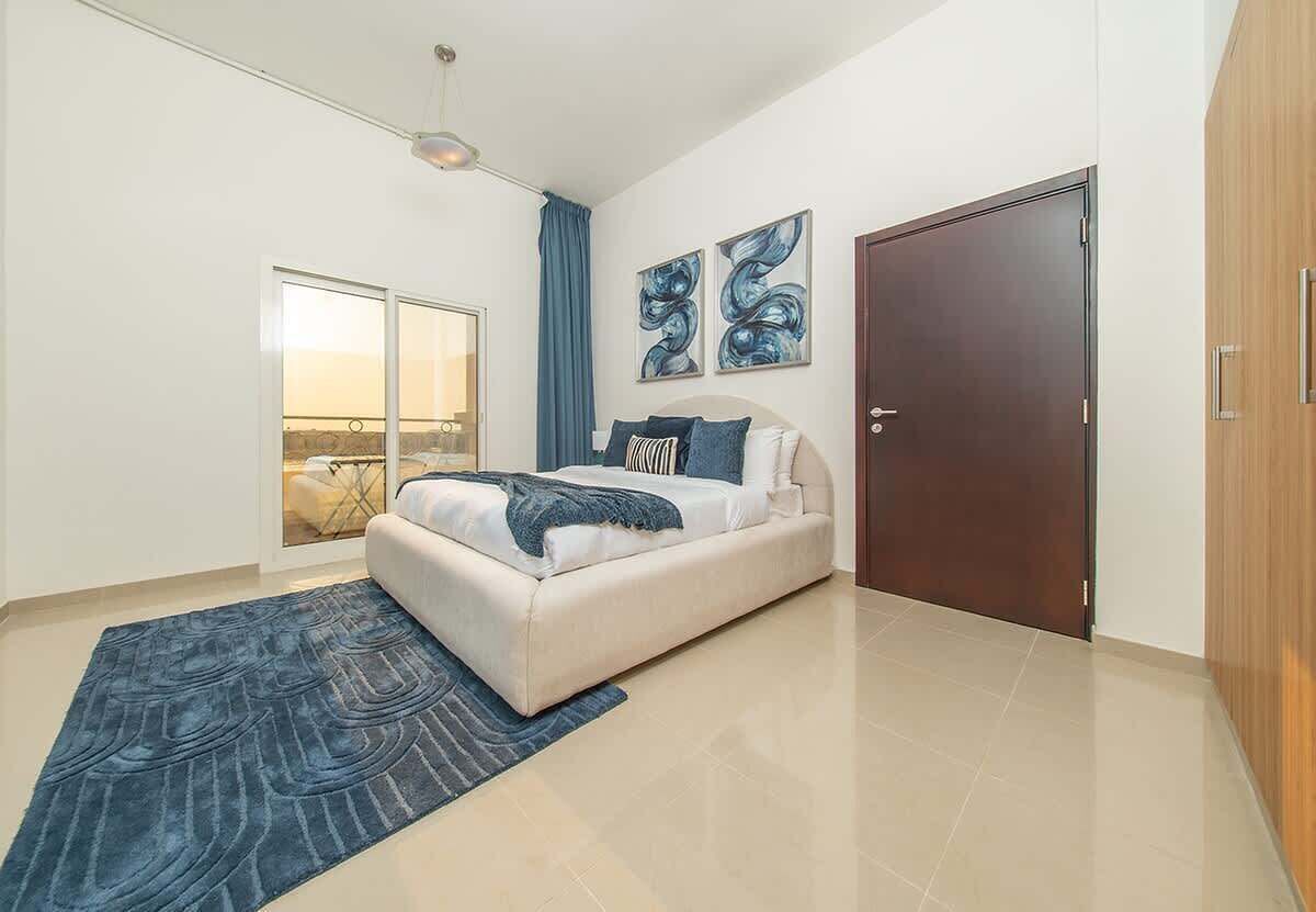A bedroom with a bed, a blue rug, a door, and a view through a glass door.