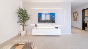 A modern living room with a large flat-screen TV mounted on a white wall, a potted plant, a white console table, and a patterned rug. - Elegant Waterfront Villa W Sunset Terrace (Ajmal Makan)