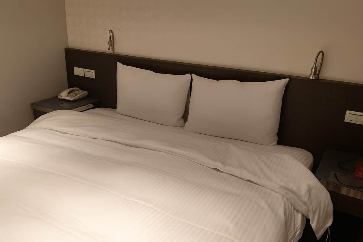 A neatly made bed with white bedding, a bedside telephone, and a nightstand with a lamp.