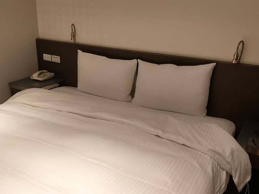 A neatly made bed with white bedding, a bedside telephone, and a nightstand with a lamp.