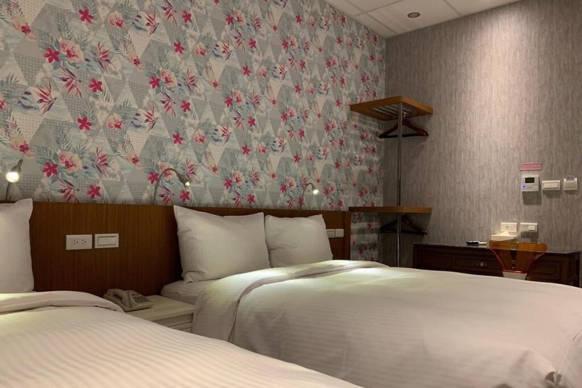 A hotel room with two beds, a wooden headboard, a floral-patterned wall, a wooden shelf, and a desk with a telephone.