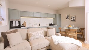 Una sala de estar moderna con un sofá color beige, una mesa de comedor de madera con sillas, y una zona de cocina con armarios azul claro y salpicadero blanco.