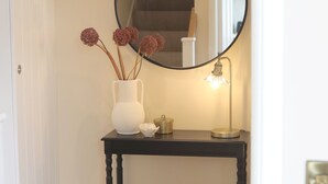 Un pasillo con un espejo redondo en la pared, un jarrón blanco con flores secas, una mesita con una lámpara y un banco de madera.