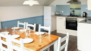 A dining area with a wooden table set for four, white chairs, a white pendant light, a white refrigerator, a stove, and a kettle. - Ocean Tide Villa 3 Hot Tub (Woolacombe)