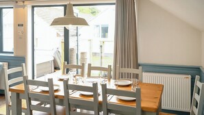 A dining area with a wooden table and white chairs, a hanging pendant light, and a view through a glass door. - Ocean Tide Villa 3 Hot Tub (Woolacombe)