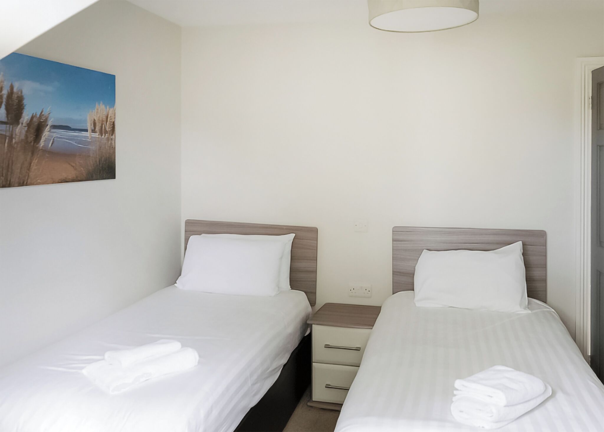 Une chambre d’hôtel avec deux lits simples, une table de chevet, un petit tiroir, un tableau au mur et un luminaire au plafond.