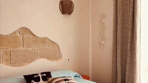 Un dormitori amb capçalera de fusta amb dibuix de rajola, llit amb roba de llit blau clar, dos almoinos i un objecte decoratiu penjat a la paret, i una finestra amb cortines.