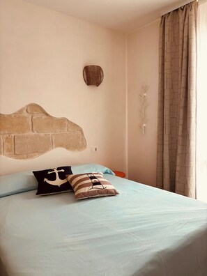 A bedroom with a brick-patterned headboard, a bed with light blue bedding, two pillows, a wall-mounted decorative item, and a window with curtains. - Queen Room with Sea View (Trapani)