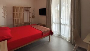 Une chambre avec un lit simple, un ventilateur de plafond, une fenêtre avec des rideaux, une télévision et une petite table.