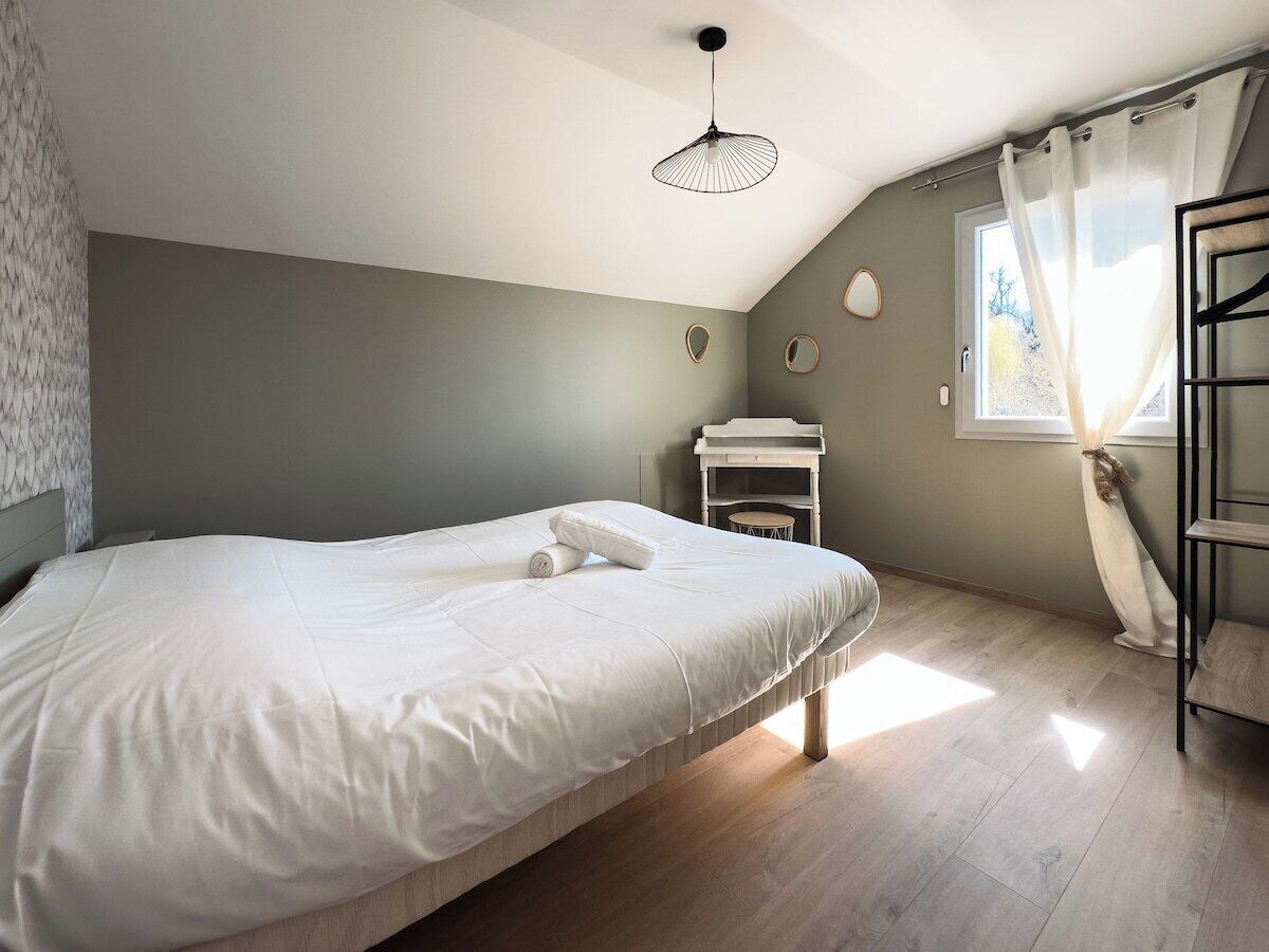 Une chambre à coucher comprenant un lit, une table de chevet, un luminaire suspendu, une fenêtre avec des rideaux et une vue sur l’extérieur.