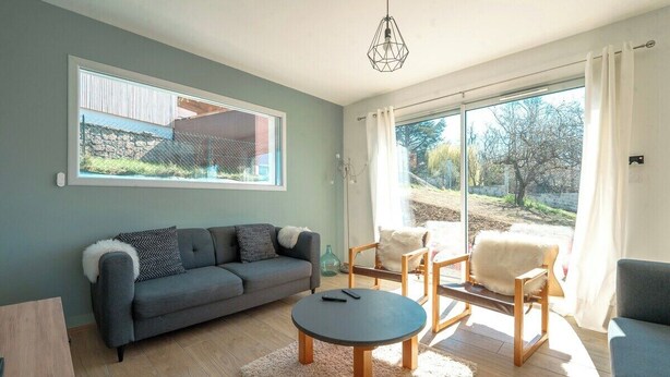 A modern living room with a sofa, a round coffee table, and a chair. There is a large window with a view of the outdoors.