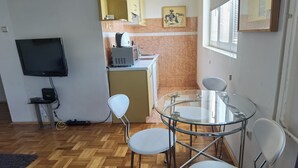 Eine kleine Küche mit Mikrowelle, Wasserkocher und Flachbildfernseher. Zwei weiße Stühle stehen um einen Glastisch.
