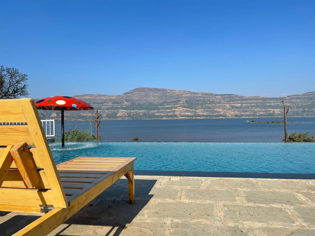 A wooden lounge chair and umbrella by a clear blue pool with a view of distant hills.