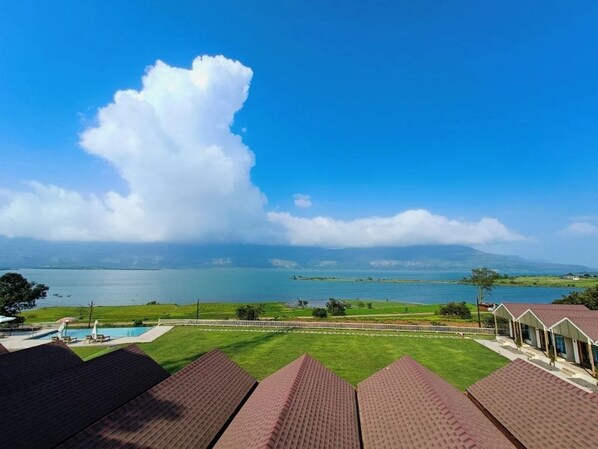 A view of a hotel with a pool, a lawn, and a large body of water in the background. - Blu Water Resort (Junnar)