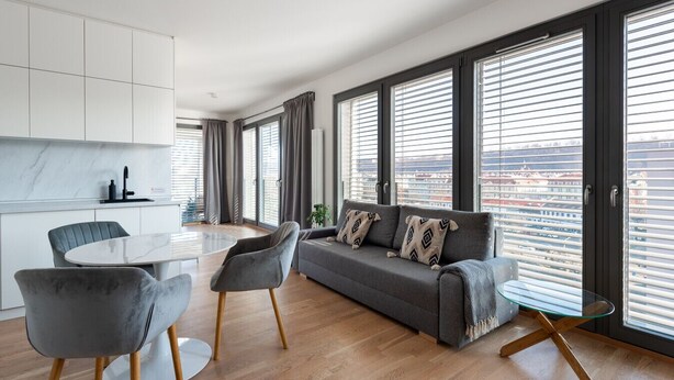 Ein modernes Wohnzimmer mit einem grauen Sofa, zwei Sesseln, einem Glastisch und einem Essbereich mit rundem Tisch. Große Fenster mit Jalousien bieten Blick auf die Stadtlandschaft.