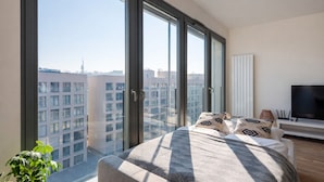 Ein modernes Schlafzimmer mit großem Fenster, einem Bett mit gemusterten Kissen, einem Fernseher und einer Pflanze.
