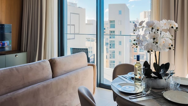 A modern hotel room with a large window, a sofa, a dining table set with plates, glasses, and a bottle of wine. - Mercury Suites (St. Julian's)