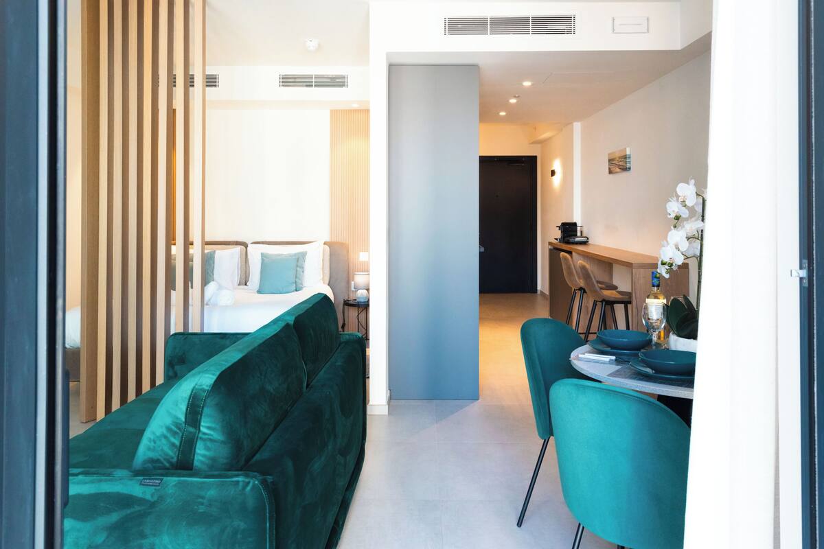 Una habitación de hotel moderna con una cama, un sofá verde, una zona de comedor con una mesa y sillas, y una cocina al fondo.