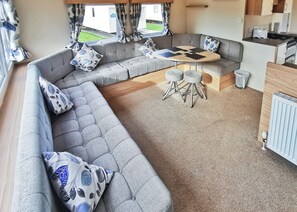A cozy RV interior with a sectional sofa, a small round table, and a kitchenette. - Gold Plus 3 Bed No Pets No Decking Sleeps 6 (Clarach Bay, Nr Aberystwyth)