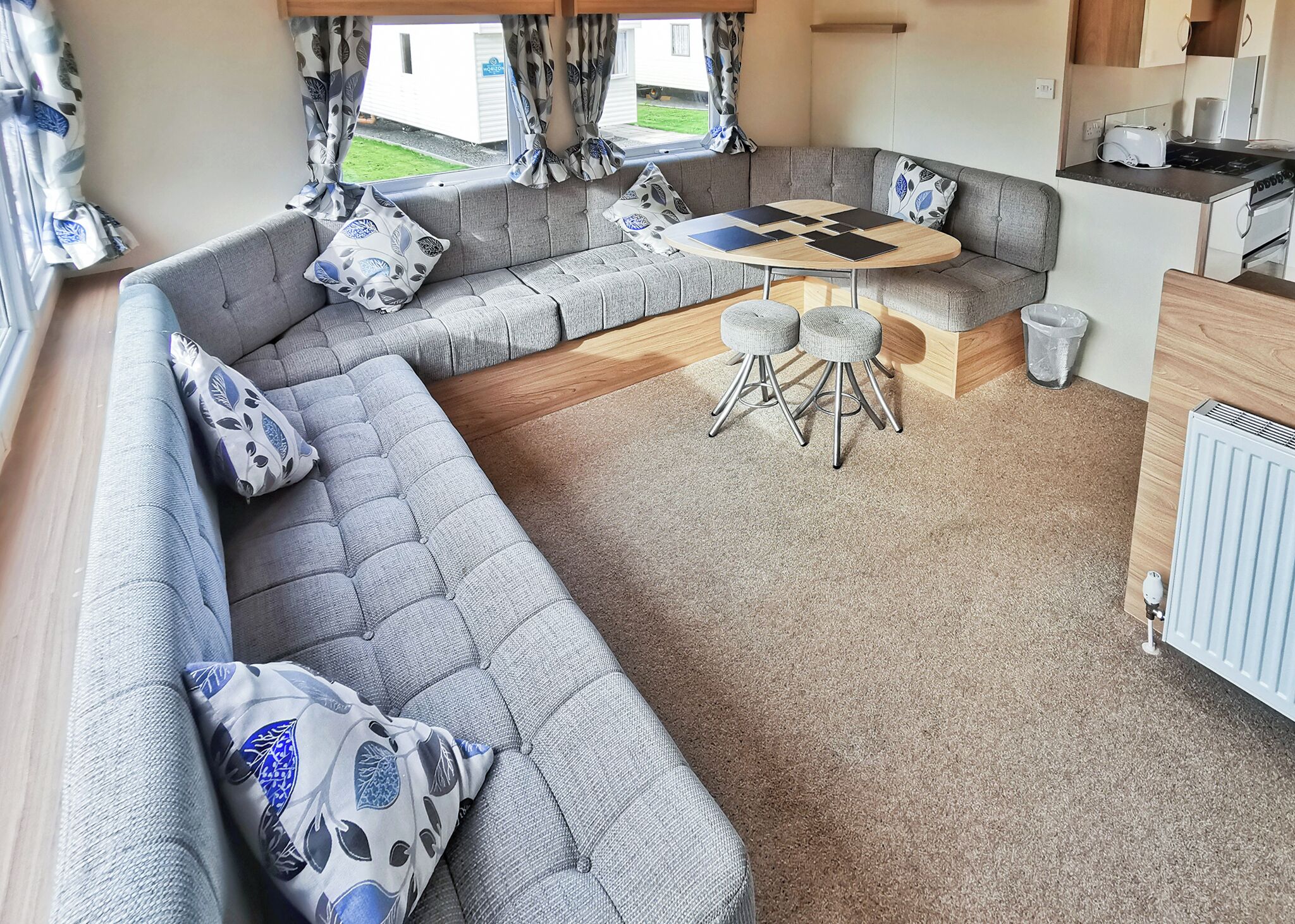 A caravan interior with a grey sofa, patterned cushions, a round table, and a kitchen area.