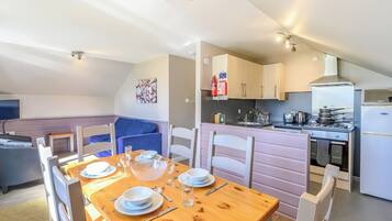 Un coin repas avec une table en bois dressée pour quatre personnes, une cuisine équipée d’appareils modernes et un espace salon confortable avec un canapé bleu.
