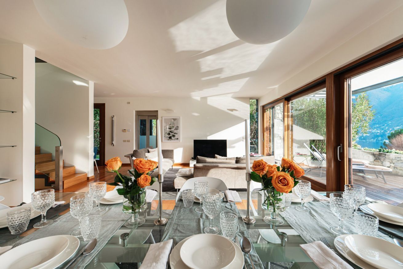 A modern dining area with a glass table set for a meal, a sofa in the background, and a view of the outdoors.