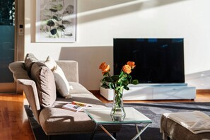 Een moderne woonkamer met een bank, een glazen salontafel, een vaas met bloemen en een flatscreen televisie.