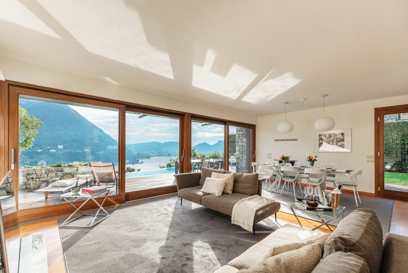 A spacious living area with a large sliding glass door leading to a balcony, a sofa, a dining table with chairs, and a view of a lake and mountains.